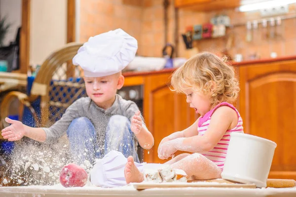 Dva sourozenci - chlapec a dívka - šéfkuchaře klobouky sedí na kuchyni. — Stock fotografie