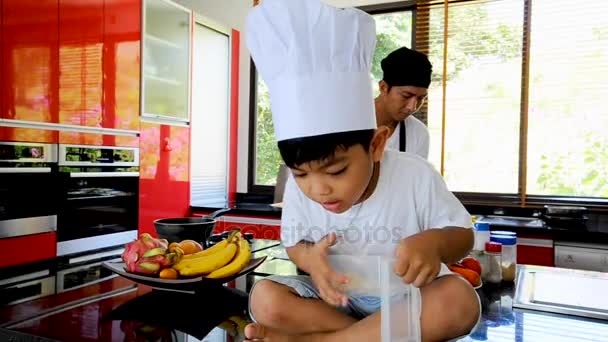 Приватні тайський шеф-кухар приготування їжі, його маленький син в капелюсі шеф-кухаря неподалік сидять на столі в сучасному стилі домашньої кухні. Дитина їсть, тато, приготування їжі — стокове відео