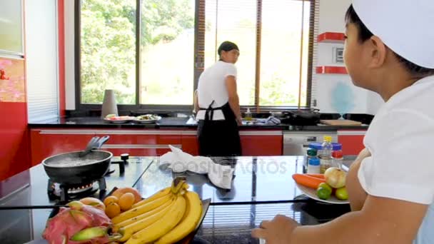 Thailändischer Koch beim Kochen, sein kleiner Sohn mit Kochmütze in der Nähe sitzt auf dem Tisch in einer modernen Wohnküche. Kind isst, Papa macht Essen — Stockvideo