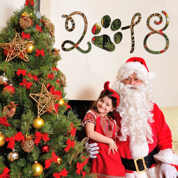 Bom espírito de Ano Novo: árvore, saco de presente grande, lareira e decoração para o ano do cão - Papai Noel e uma linda vestida com traje vermelho menina com pequenos chifres vermelhos sentados perto dele — Fotografia de Stock