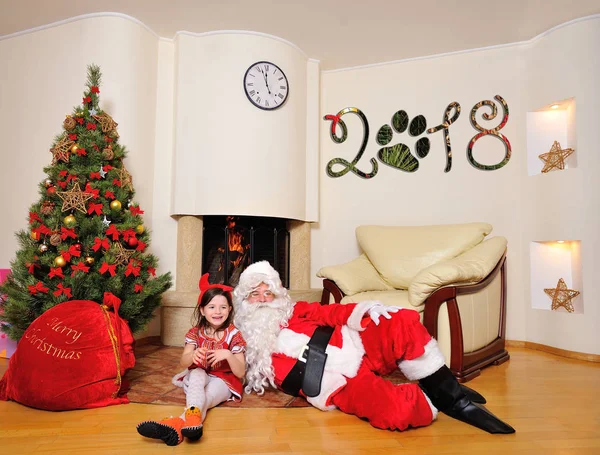 Bom espírito de Ano Novo: árvore, saco de presente grande, lareira e decoração para o ano do cão - Papai Noel deitar no chão e um bonito vestido de menina traje vermelho com pequenos chifres vermelhos sentados perto dele — Fotografia de Stock