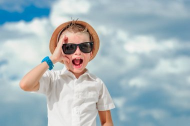 Portre bulutlar bir arka plan üzerinde yakından: yakışıklı küçük çocuk güneş gözlüğü ve fedora şapkası ile ince uygun gömlek giymiş, o elini yüzünü ve çığlıklar içinde için kaldırdı