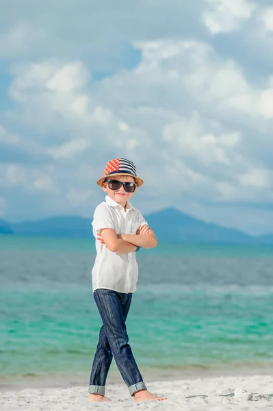 Volle groei portret op een tropisch strand: dappere knappe 8 jaar oude jongen in solide heldere slim fit overhemd, donkere broek met zonnebril en fedora hoed, permanent barefoot op zand — Stockfoto