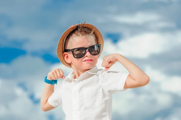 Blisko do portretu na tle chmur: przystojny chłopiec ubrany w stałych jasnej koszuli okulary przeciwsłoneczne i kapelusz fedora. Pochylił kolana, pokazuje jak silny jest. Patrząc w dal — Zdjęcie stockowe