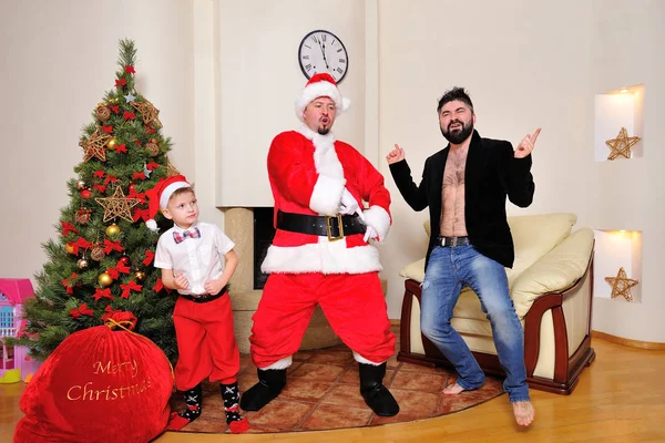Christmas tree, gift bag, fireplace - men dressed as Santa, boy in red pants and high socks and a hipster in velours jacket on bare torso dancing, play the fool. Looking crazy