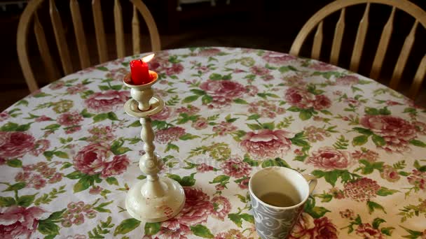 Tazza di tè e candela accesa su un tavolo. Sedie di legno. Tovaglia con fiori. Sala interna . — Video Stock