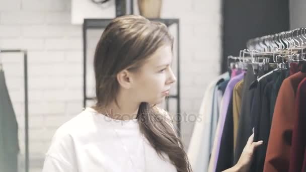 Ung kvinna i en T shirt är att välja kläder i en butik — Stockvideo