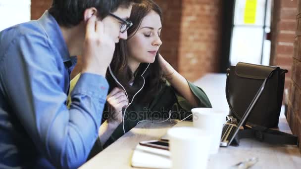 Vista lateral de um casal milenar usando fones de ouvido juntos em um café — Vídeo de Stock