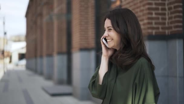 Милая девушка в зеленой рубашке разговаривает по телефону на улице, вид сбоку — стоковое видео