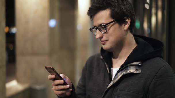 Joven en gafas es mensajes de texto en la calle en la vista lateral de la noche — Vídeos de Stock