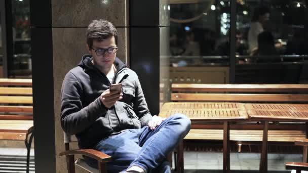 Joven sentado en un café de la calle por la noche con un teléfono inteligente — Vídeo de stock