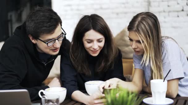 Três amigos estão assistindo a um vídeo em um smartphone em um café ao ar livre — Vídeo de Stock