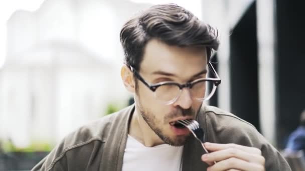 Hipster guy eating meat and talking to a friend in a street cafe, front view — Stock Video