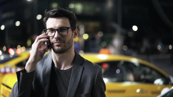 Junger Geschäftsmann mit Brille am Telefon, im Hintergrund ein Taxi — Stockvideo