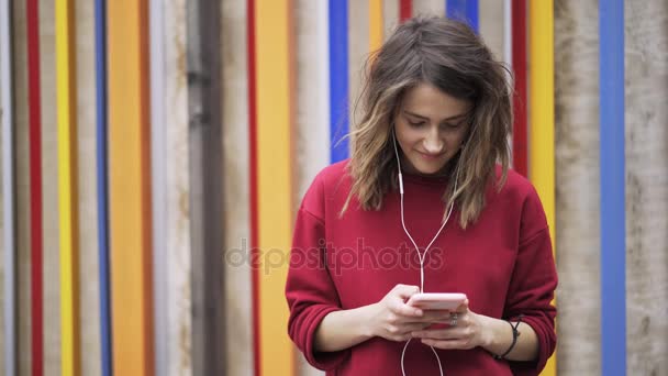 Молода жінка в червоному смс і прослуховування музики біля барвистої стіни, крупним планом — стокове відео