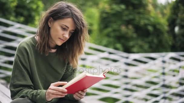 Joven estudiante universitario en verde leyendo un libro en un parque — Vídeo de stock