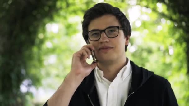 Молодой человек в очках разговаривает по телефону и гуляет в парке — стоковое видео