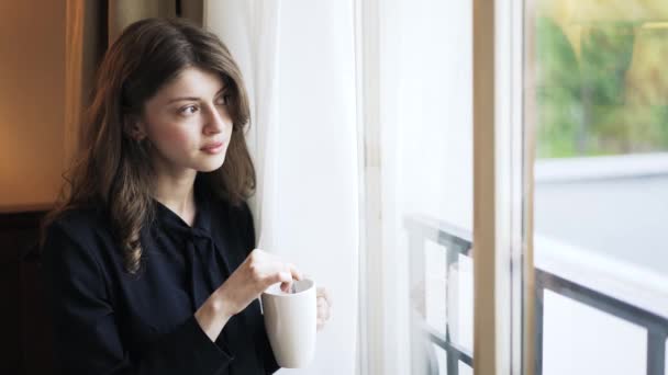 Молода жінка з чашкою дивиться крізь вікно — стокове відео