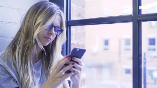 Blond tonåring flicka webbsurfning med hennes smartphone på en fönsterbräda — Stockvideo