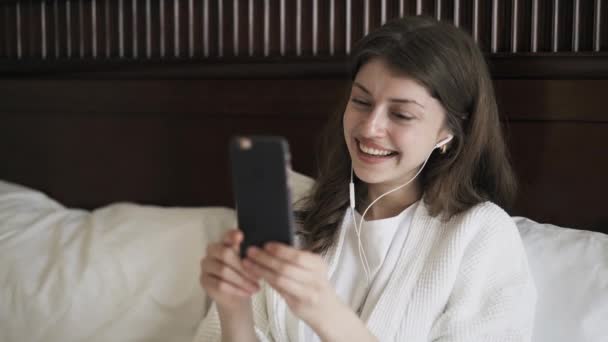 Молодая женщина делает видео-звонок со своим смартфоном в постели, закрыть — стоковое видео