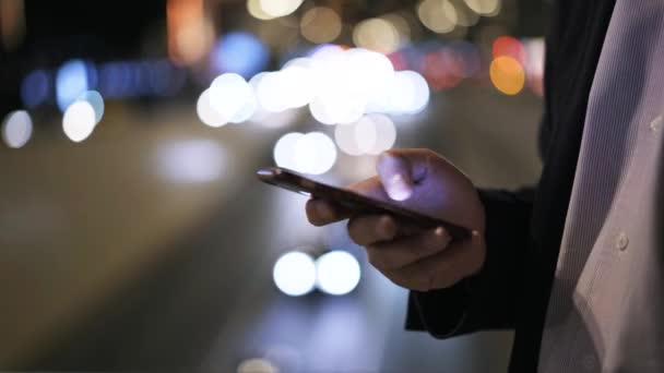 Vista lateral de um homem de terno web surf com um smartphone perto de uma estrada noturna da cidade — Vídeo de Stock