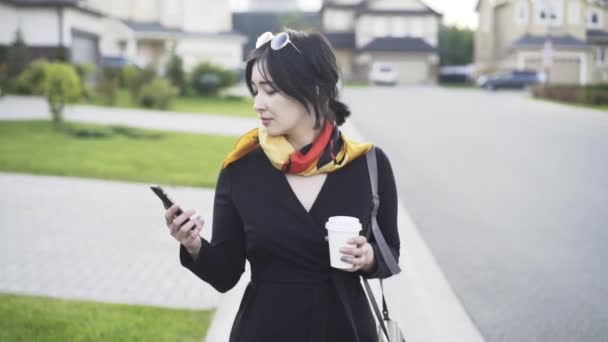 Улыбающаяся азиатка с кофе смотрит на телефон и ходит — стоковое видео