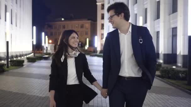 Νεαρό ζευγάρι περπάτημα σε μια πόλη νύχτα, κρατώντας τα χέρια και να χαμογελά επιχειρηματίες — Αρχείο Βίντεο