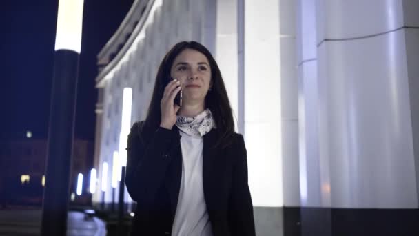 Mujer de negocios caminando en una ciudad nocturna y hablando en su teléfono inteligente — Vídeos de Stock