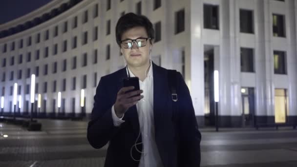 Молодий бізнесмен зі смартфоном і навушниками гуляє в нічному місті — стокове відео