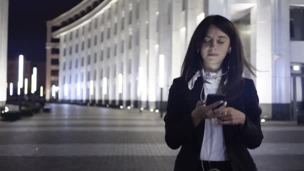 Mujer joven de cabello oscuro con un teléfono inteligente y auriculares camina en una ciudad nocturna — Vídeos de Stock