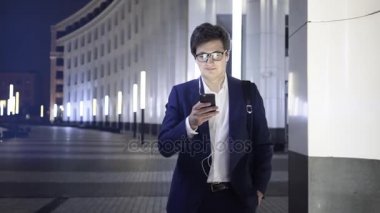 Bir akıllı telefon ve kulaklık ile genç işadamı gece şehir sokak yürür