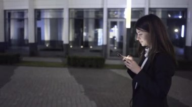 Bir akıllı telefon ile genç iş kadını bir gece şehirde sokak, yan görünüm yürür