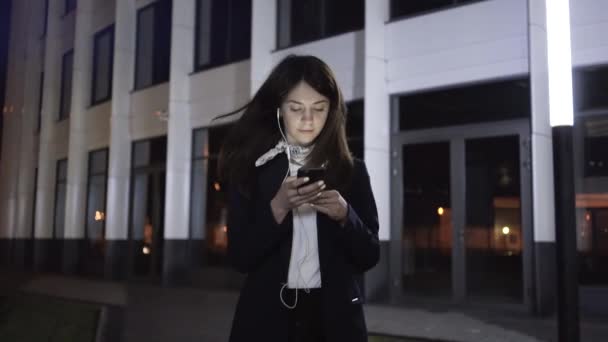 Монтаж молодой бизнесвумен со смартфоном и наушниками в ночном городе — стоковое видео