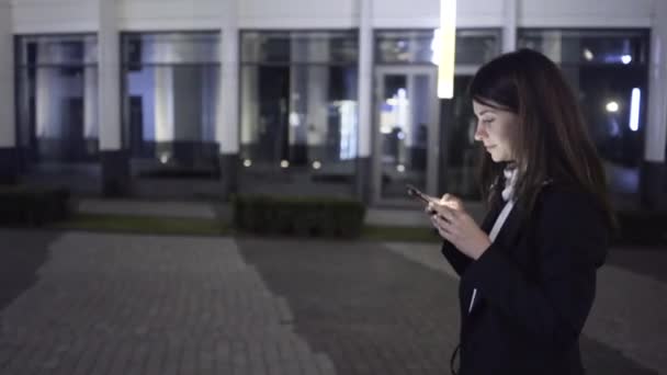 Joven mujer de negocios con un teléfono inteligente camina en una calle nocturna de la ciudad, vista lateral — Vídeos de Stock