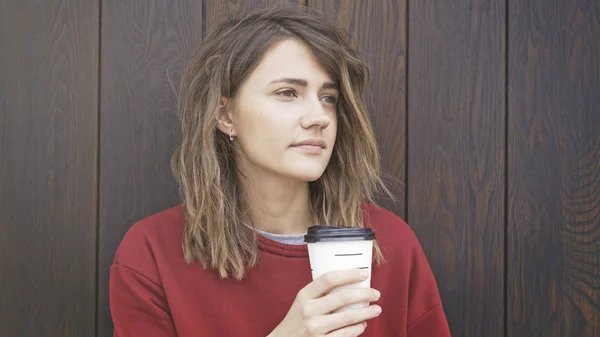Retrato de una mujer joven con un suéter rojo está tomando café para ir de pie cerca de una pared de madera. Tiro medio — Foto de Stock