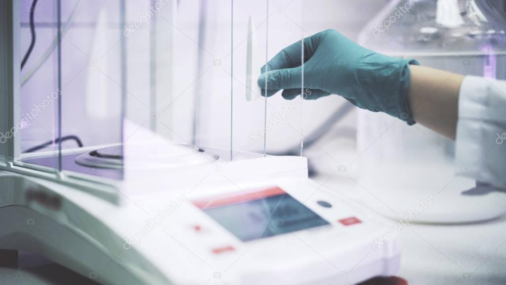 Scientist s hand in a green glove is putting liquid at electronic scales in a lab. Medium shot