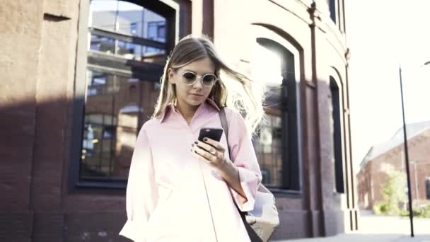 Mujer rubia en gafas de sol caminando y hablando en su smartphone — Vídeos de Stock