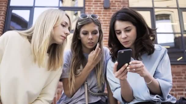 Três mulheres olhando para uma tela de smartphone sentado no verão rua zoom out — Vídeo de Stock