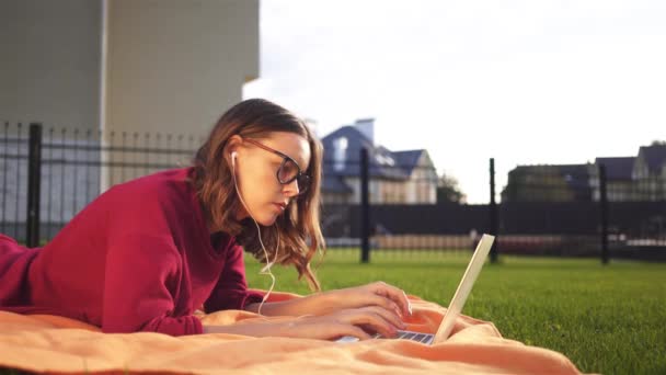 Geconcentreerd hipster meisje in glazen liggen in haar achtertuin met een laptop — Stockvideo