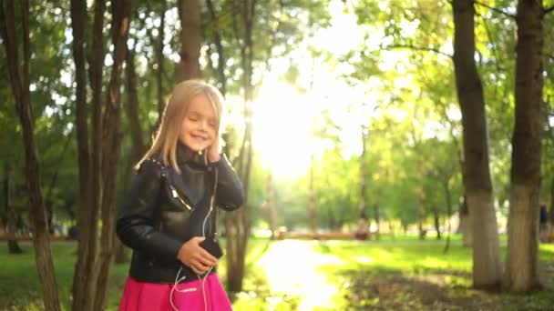 Маленькая девочка в кожаной куртке слушает музыку в парке — стоковое видео