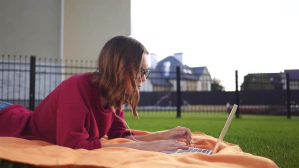 Hipster chica acostada en la hierba con un ordenador portátil, lado — Vídeos de Stock