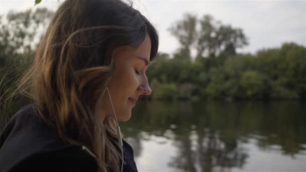 Вид збоку жінки, що слухає музику і йде вздовж річки — стокове відео