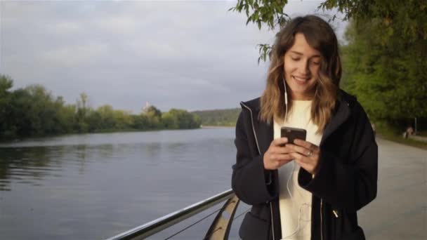 Mujer joven tranquila escuchando música y caminando cerca de un río — Vídeo de stock