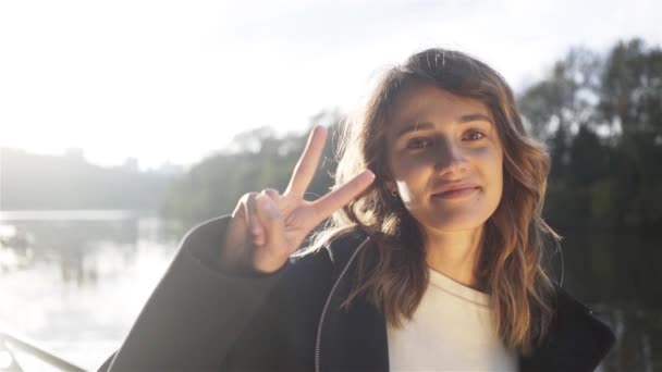 Mooie jonge vrouw toont vredesteken vlakbij een rivier — Stockvideo