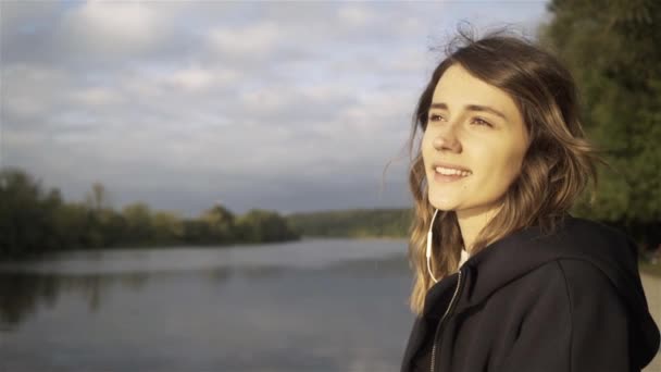 Весела молода жінка слухає музику і стоїть на березі річки — стокове відео