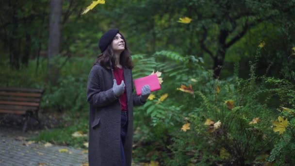 Heureuse femme brune avec un livre souriant marchant sous un feuillage — Video