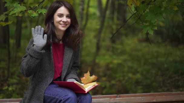 Усміхнена брюнетка з книгою махає рукою в осінньому парку — стокове відео