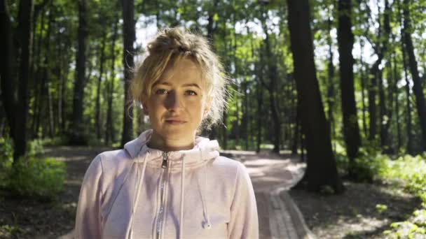 Jeune femme en sweat-shirt rose regardant la caméra et souriant dans un parc — Video