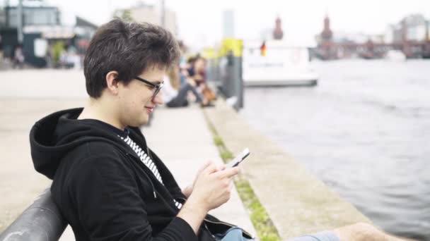 Joven en pantalones cortos enviando mensajes de texto y sonriendo cerca del puente Oberbaum en Alemania — Vídeos de Stock