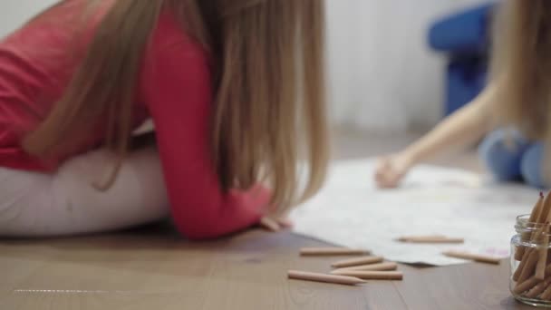 Две маленькие девочки рисуют большую картинку на полу карандашами — стоковое видео